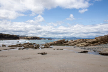 Scenic coastal landscape with sandy beach, unique rock formations, and ocean waves under a partly cloudy sky. Distant mountains enhance the natural beauty