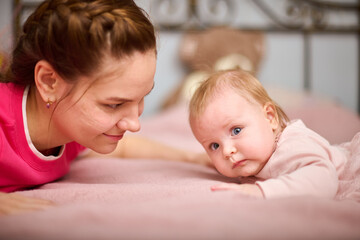 Young mother with brown hair and baby girl lying on pink blanket. Warm lighting, indoor setting, expressions of joy and warmth, capturing tender bonding moment