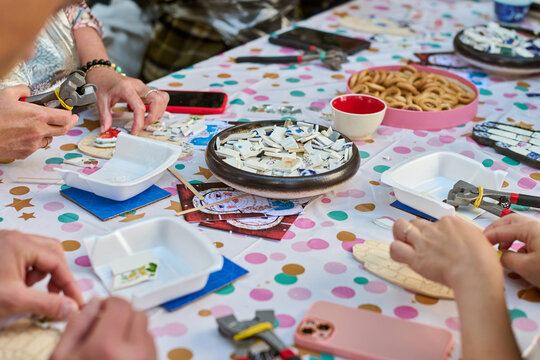 Hands piece together mosaic art with ceramic tiles. Colorful table setting, scattered materials. Bright ambient lighting enhances intricate details