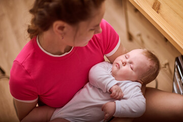 Young woman with blonde hair cradles infant in pink room. Baby girl sleeps peacefully in woman lap. Gentle lighting and warm ambiance enhance soothing scene