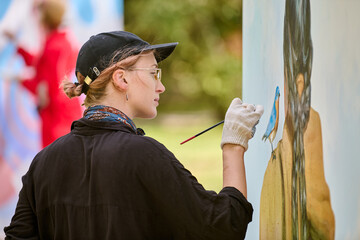 Young adult, blonde, caucasian woman paints outdoors. Wearing glasses, cap, focused on canvas....