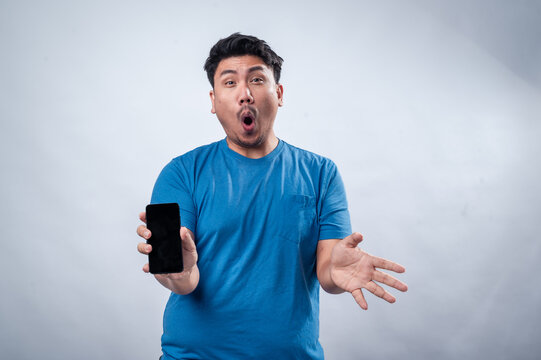 An Asian man in a blue t-shirt holds a smartphone forward with an excited and surprised facial expression. His mouth is open, and he appears amazed. The background is plain white