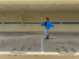 A woman in a blue jacket is dancing in a parking lot. Scene is lighthearted and fun