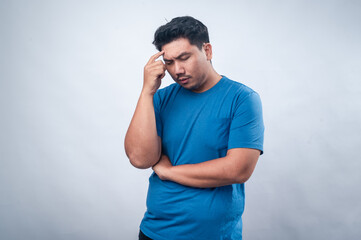 An Asian man in a blue t-shirt stands against a plain white background, pointing to his temple with a skeptical and questioning expression. His posture suggests deep thought, confusion, or curiosity