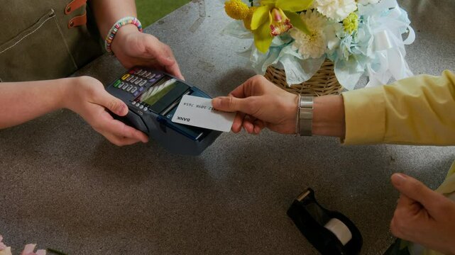 High angle view of unknown female customer buying basket of flowers and tapping card on payment terminal held by florist at counter
