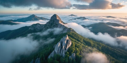 Aerial view of rugged mountain peaks emerging from dense fog with lush green forests under a dramatic sky at sunrise