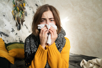 Sick young woman with a runny nose using a handkerchief at home looking sad and unwell, sitting in...