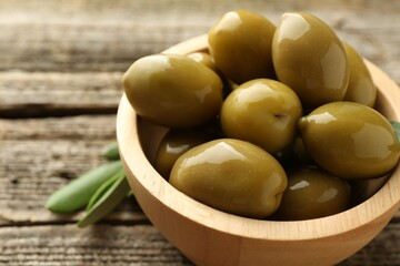 Delicious marinated olives in bowl and green leaves on wooden table, closeup