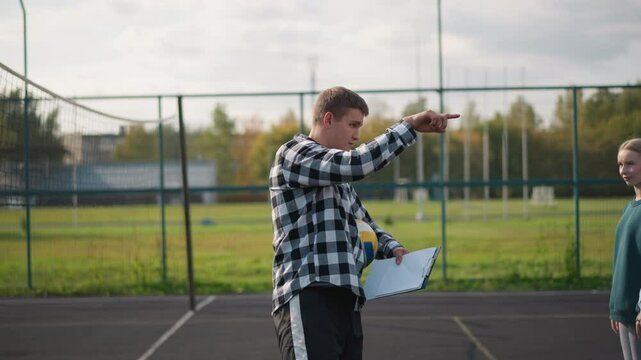 Volleyball coach with ball under arm and record book instructing female students to walk around court during practice session in outdoor environment, giving directions to beginners