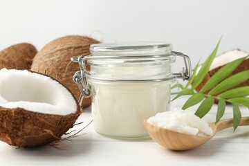 Organic coconut cooking oil in jar, spoon, fresh fruits and green branch on white wooden table, closeup