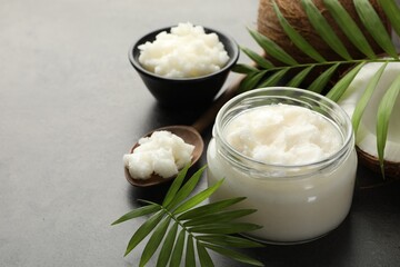 Organic coconut cooking oil in jar, spoon, fresh fruits and green branches on grey table, closeup....