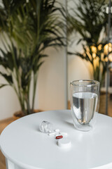 White and red pills and capsules spilling from an open pill bottle next to a glass of clean drinking water on a white side table, with a blurred palm tree background creating a calm atmosphere