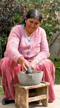 vertical old woman of the aymara chola culture grinding in a mortar or also called batan in the courtyard of her house in la paz, bolivia - cooking concept