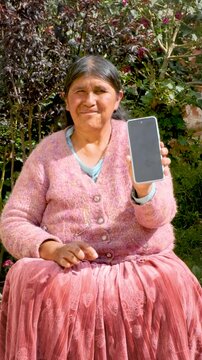 vertical cholita adult woman in pollera showing her cell phone to the camera in the courtyard of her house in La Paz Bolivia - concept of older adults