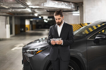 Young Caucasian male businessman counts cash in underground parking lot next to car, conveying...