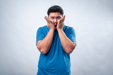 An Asian man in a blue t-shirt stands against a white background, expressing fear or anxiety with...