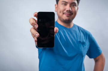 An Asian man wearing a blue t-shirt holds a smartphone in one hand and points at it with the other while smiling confidently. He is posing against a plain white isolated background