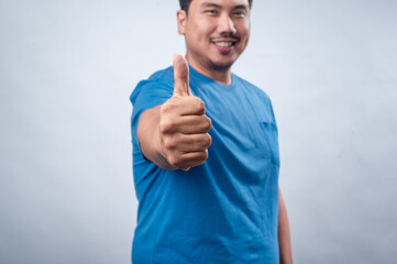 A cheerful Asian man wearing a blue t-shirt gives a thumbs-up gesture while smiling, standing...