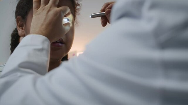 Male doctor in white coat examining eyes of little Black girl using diagnostic penlight checking pupil reflex at doctors appointment