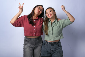 Obraz premium Two young Asian women with long hair wear casual crop shirts, one red and one green, with dark jeans. They make playful faces and peace signs while posing together against a plain white background.