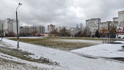 Behind a metal fence, surrounded by trees and buildings, is a stadium with a grass soccer field with goals, running tracks, and poles with lights. It snowed in the winter. Cloudy weather