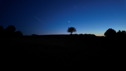 Countryside silhouettes under the stars, meteor trail and moonlight.