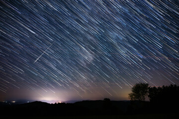 Milky Way stars with meteor shower trails and countryside silhouettes.