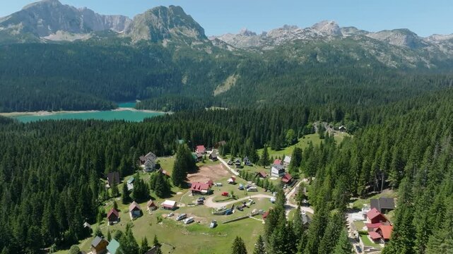 Cinematic drone pullback from a small camp village in a pine forest on the shore of the blue Black Lake, revealing the Durmitor mountain range of the Dinaric Alps in summer
