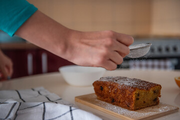 Confectioner sprinkles powdered sugar over a freshly baked loaf cake on a wooden board in a cozy kitchen. The sweet treat is ready to be served. Homemade cooking
