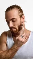 Fototapeta premium Man with beard gently holding glass skincare bottle to cheek, showcasing smooth skin and hydration against white studio background. Concept of male beauty, skin care products promotion