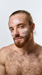 Obraz premium Portrait of handsome bearded shirtless man gazing confidently with white face cream applied across forehead and cheeks, promoting balanced skincare. Concept of male beauty, skin care products
