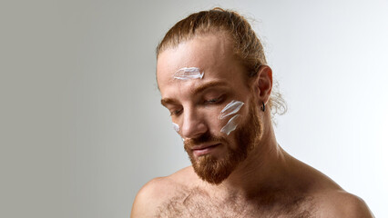 Man with tied-back hair and beard looking downward with white face cream applied, expressing calmness and skincare awareness against white studio background. Concept of male beauty, skin care