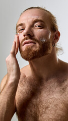 Obraz premium Portrait of handsome man with smooth skin and groomed beard applying cream to face against white studio background. Concept of male beauty, skin care products, healthy skin