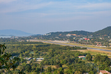 Obraz premium Panoramic view from Kao Hua Jook Pagoda of Ko Samui Airport with several aircraft on the runway, surrounded by lush tropical forest, hillside developments, and coastal mountains in the misty distance