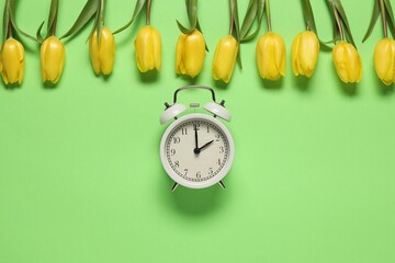 Spring time. Alarm clock and beautiful tulips on green background, flat lay