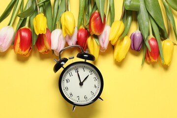 Spring time. Alarm clock and beautiful tulips on yellow background, flat lay
