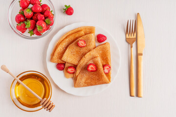 Top view of traditional sweet folded crepe french dessert or sweet blini tasty snack made from batter served on plate with red strawberries on white wooden table with bowl of honey, fork and knife