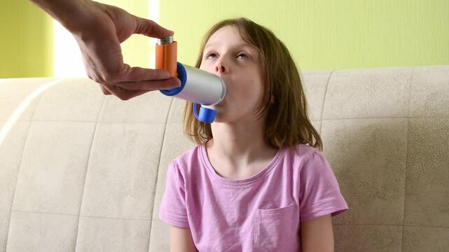 Girl sitting on couch using inhaler with spacer while adult assists. Pediatric asthma treatment, respiratory therapy, home healthcare.