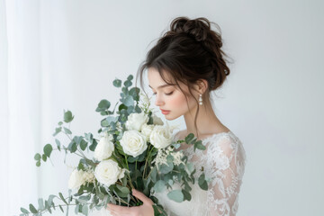 Elegant bride holding a lush bouquet of white roses and greenery in a soft, bright setting during a wedding preparation