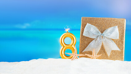 Golden glittering gift box with sparkle bow sits on sand, beach with starfish and number 8 candle with flame. Background sea creating festive atmosphere.  Birthday, International Women's Day, holiday