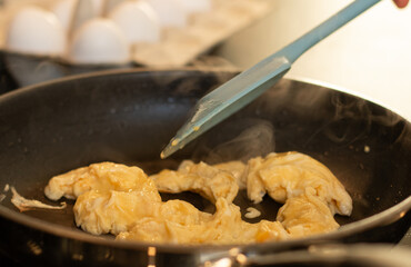 Close up of making scrambled omelet egg in a pan, blurred kitchen table background with white eggs