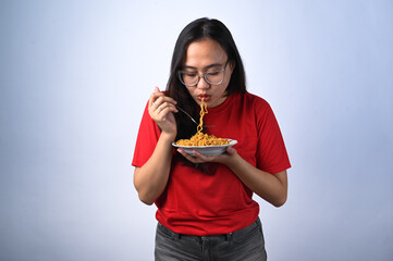 Asian woman in a red shirt and glasses enthusiastically eating instant noodles with a fork while...