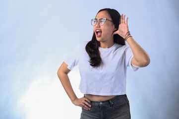 Fototapeta premium Asian woman with long dark hair, wearing glasses, a white crop top, and black jeans, cupping her hand to her ear while smiling. Studio shot with a plain white background, expressing curiosity 