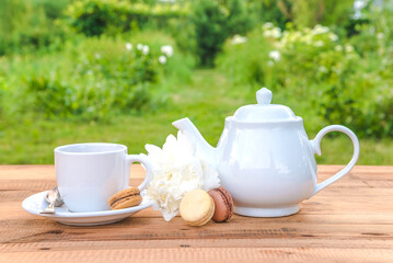 White cup of tea, tea pot, white peony flower and macaron on a wooden table in the green garden