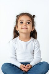 A young girl with long hair is sitting on the floor and looking up at the camera. She is wearing a white shirt and blue jeans. Concept of innocence and curiosity