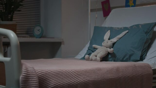No people shot of children bed in empty hospital ward with tucked cozy blanket, adorable soft bunny toy on pillow and colorful children drawings on wall