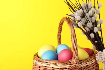 Beautiful decorated Easter eggs in wicker basket and willow branches on yellow background, closeup....