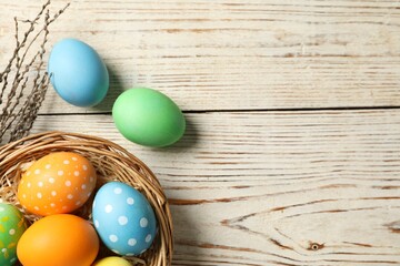 Beautiful decorated Easter eggs, wicker bowl and willow branches on light wooden table, flat lay. Space for text