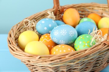 Many decorated Easter eggs in wicker basket on light blue table, closeup