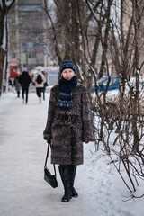 A woman in a stylish fur coat stands on the sidewalk in a snow-covered winter city, exuding elegance and charm.